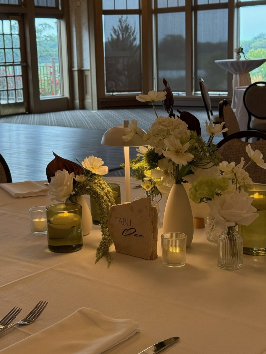 A white reception table with white blooms in bud vases, a cordless lamp and candlelight beside tall windows