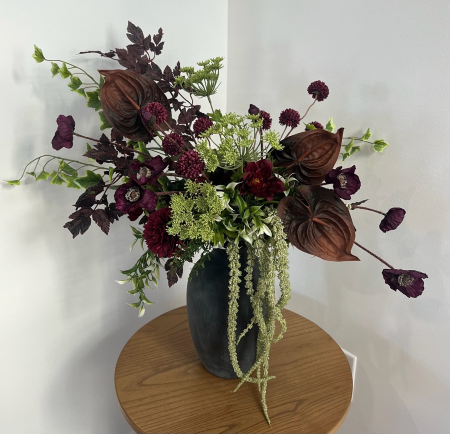 Premium fall arrangement: a fuller arrangement of chocolate anthuriums, burgundy blooms and green hydrangea with trailing amaranthus in a dark stone vase